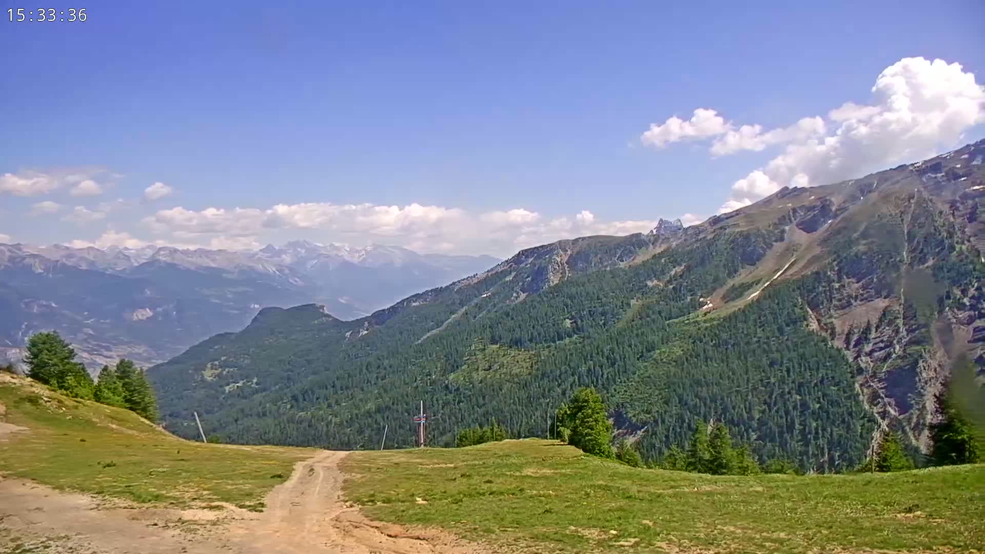 A grassy mountaintop with a dirt road overlooks a valley of evergreen forests and distant, snow-capped mountains under a mostly sunny sky with scattered clouds.