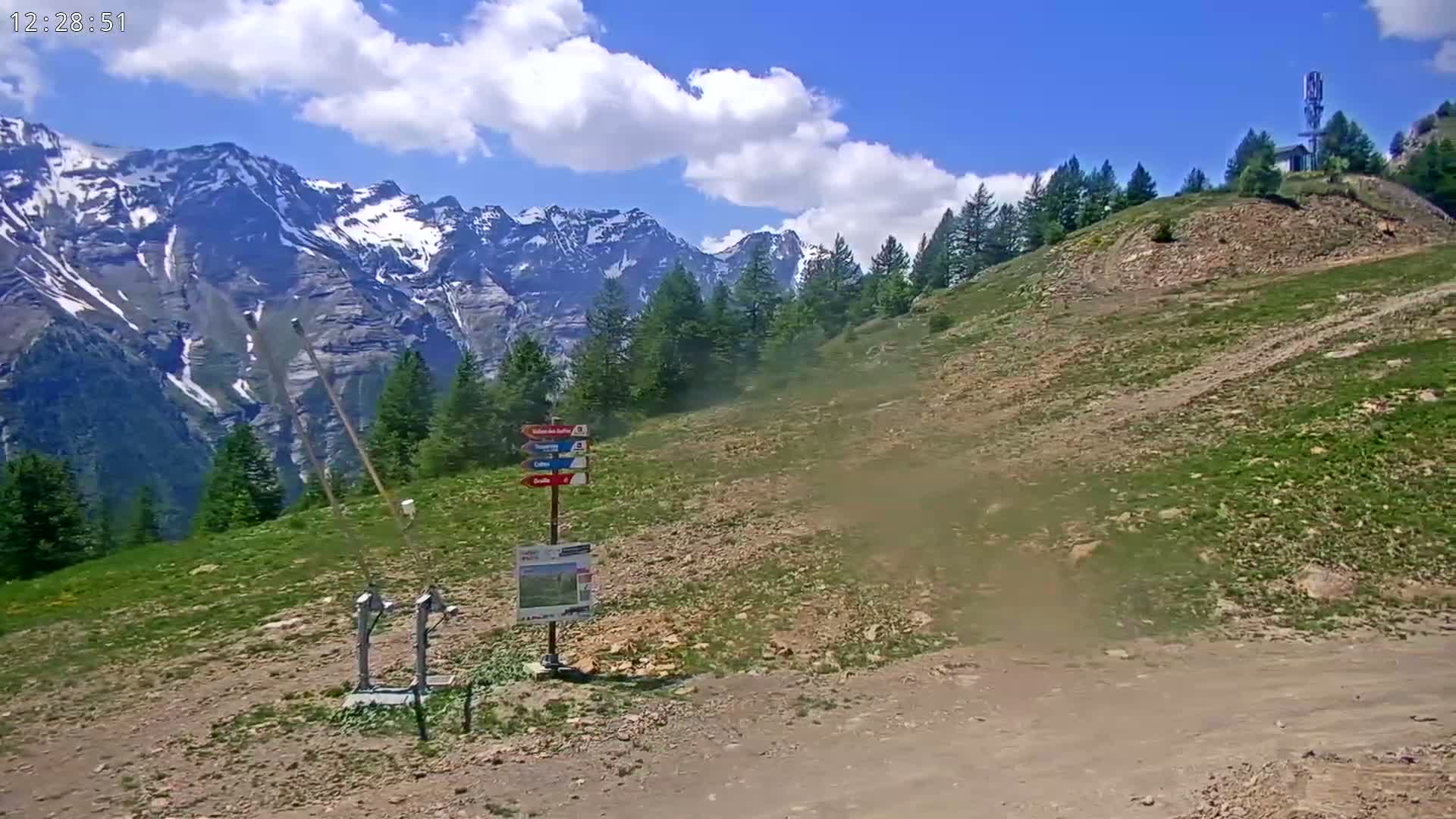 A mostly sunny mountainside scene shows a sparsely vegetated hill leading to a snow-capped mountain range under a partly cloudy sky.