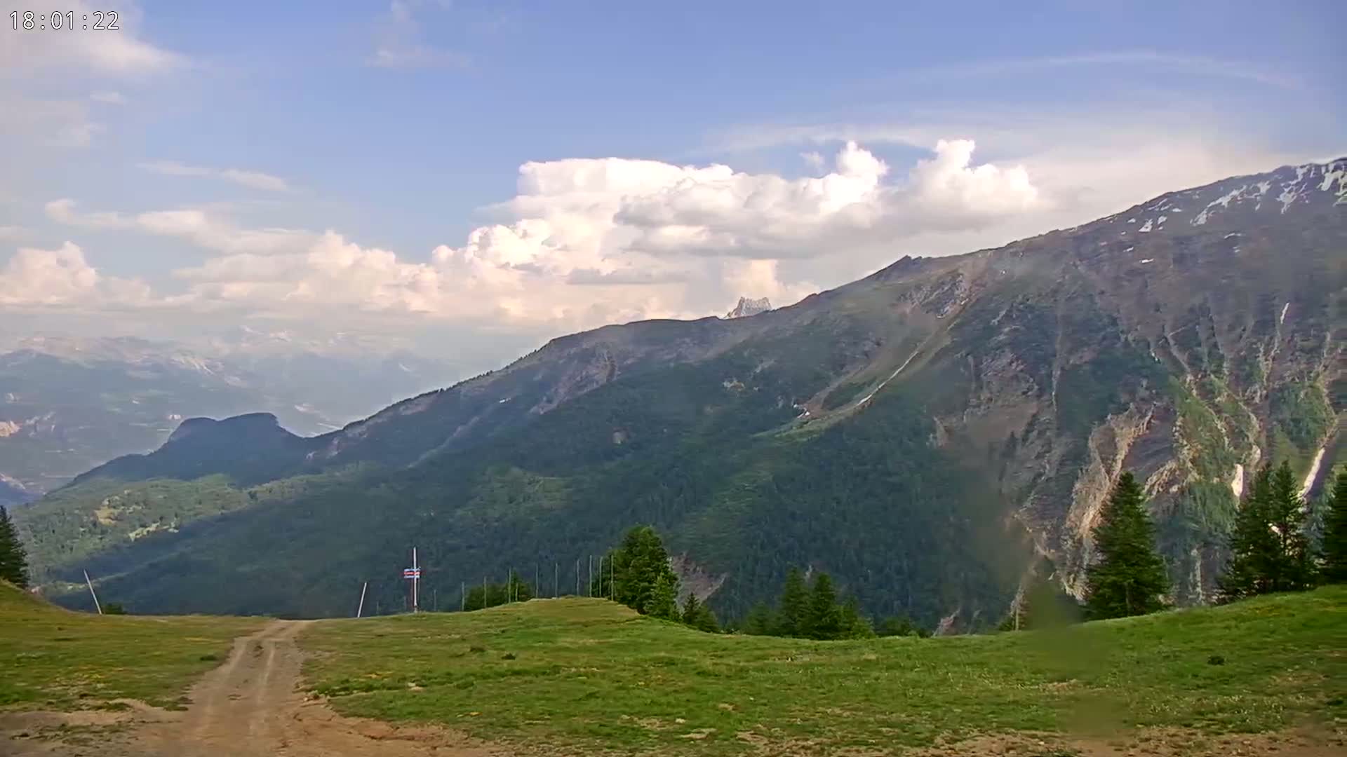 A dirt road leads to a grassy mountaintop overlooking a partly cloudy sky and a vast, verdant mountain range.