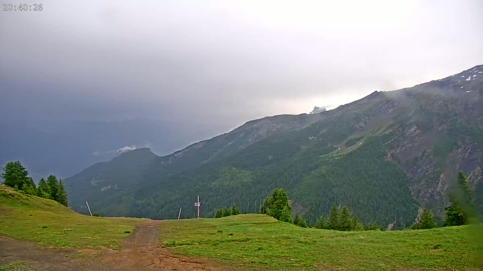A grassy mountaintop plateau overlooks a range of forested mountains under a cloudy sky.