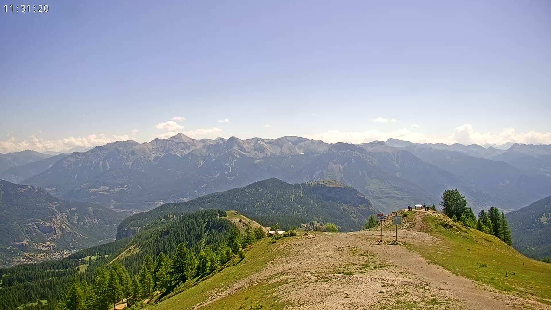A mountaintop view on a mostly clear day shows a rocky path leading to a small plateau with a few structures, overlooking a valley of evergreen trees and distant mountains under a light blue sky with a few clouds.
