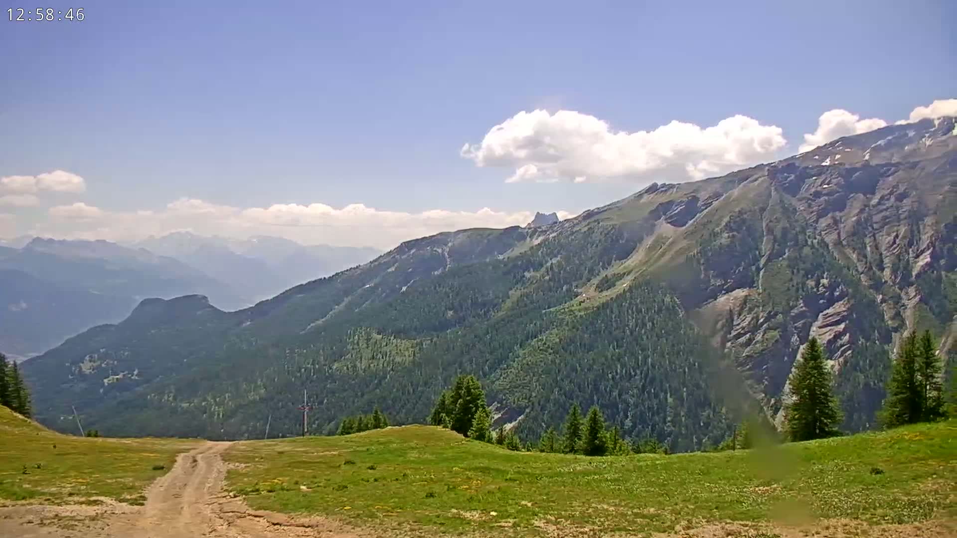 A partly cloudy sky overlooks a mountain range with a grassy foreground and a dirt road leading into the mountains.