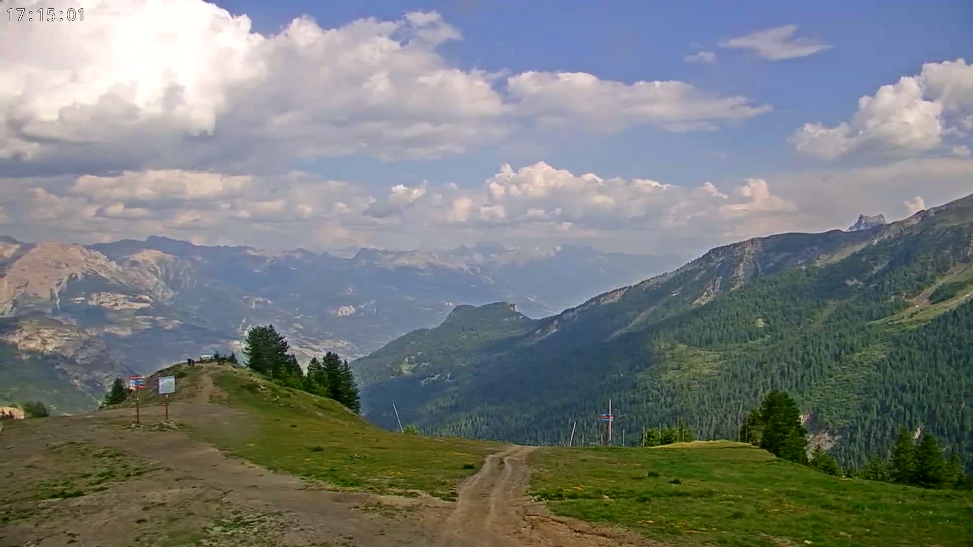 A partly cloudy sky overlooks a mountainous landscape with a grassy hilltop featuring a dirt path and a few people in the distance.