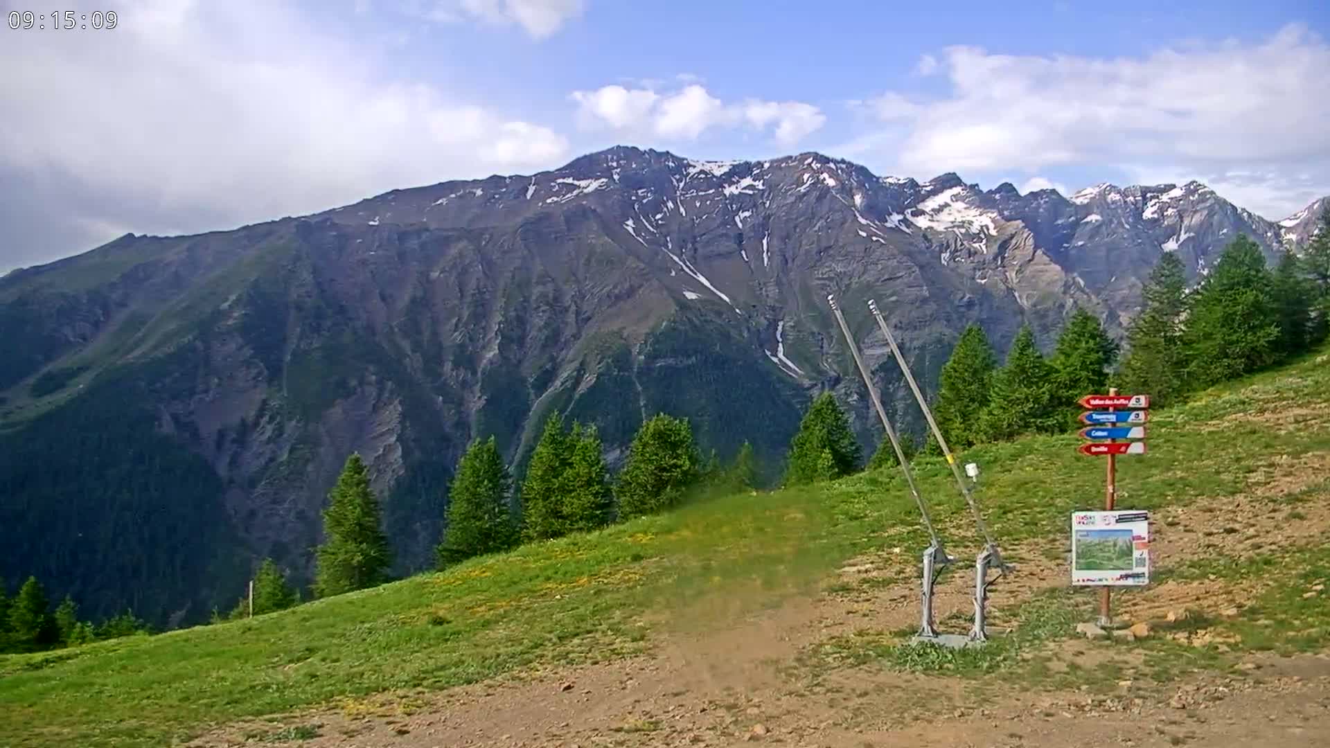 A partly cloudy day reveals a mountain range with some snow patches, viewed from a grassy hillside with a few evergreen trees and a trail marker.