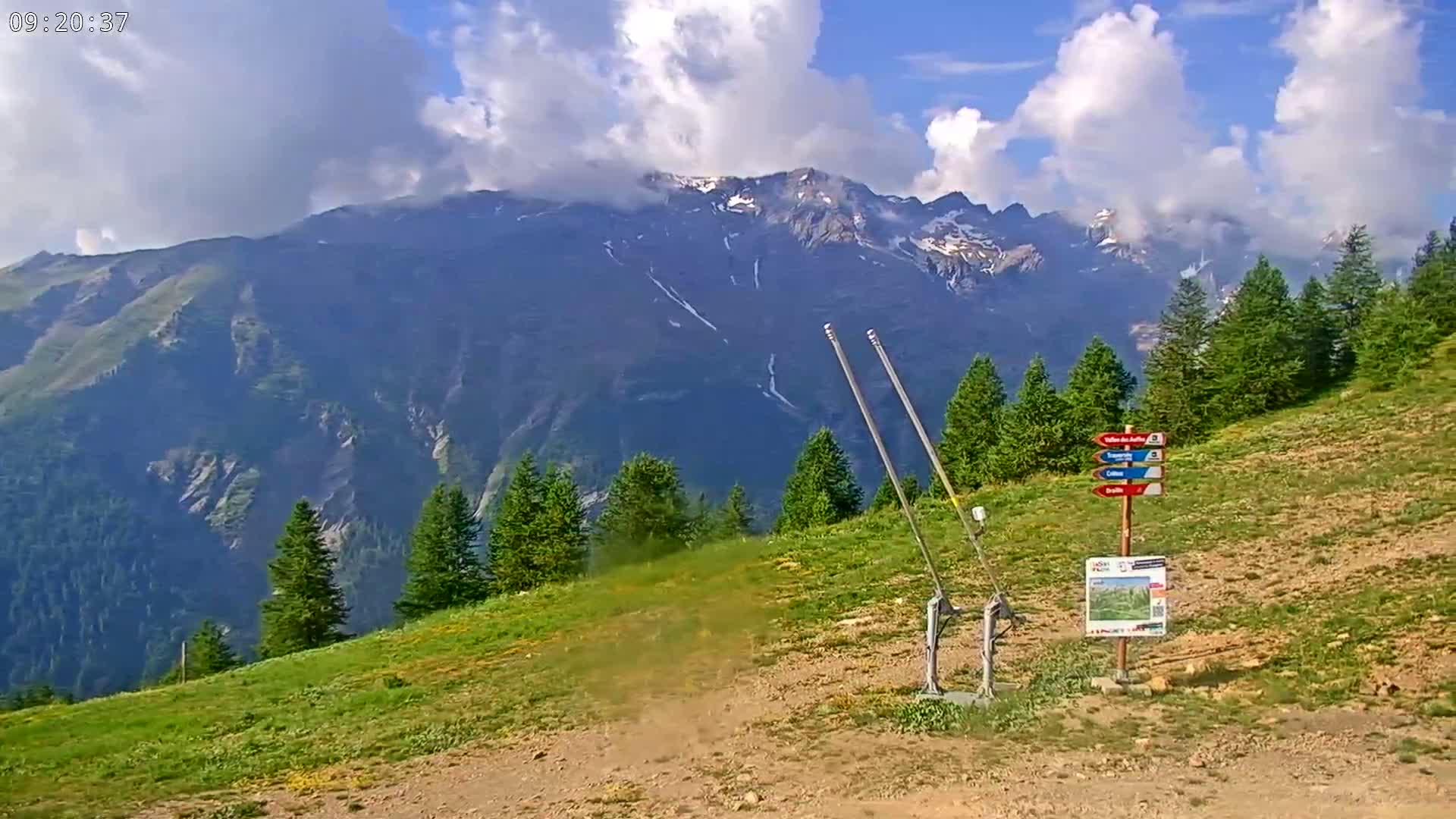 A partly cloudy day reveals a mountainous landscape with green hills, evergreen trees, and a ski lift mechanism.