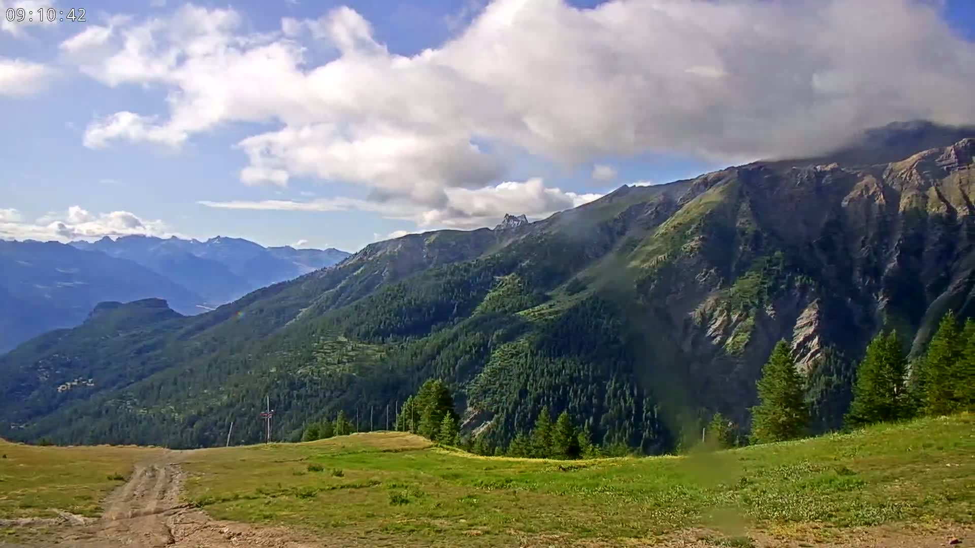 A partly cloudy day reveals a view of rolling green hills and mountains covered in coniferous trees.
