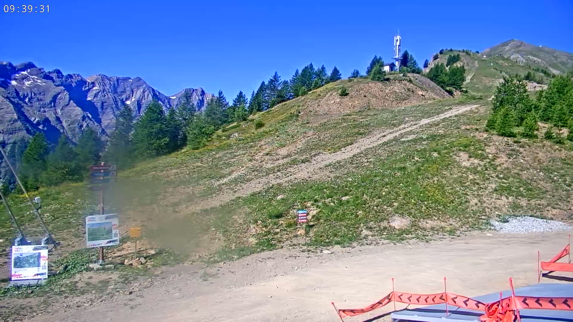 A mountainous landscape under a clear blue sky shows a dirt path leading up a hill sparsely covered in low-lying vegetation, towards a communications tower on a peak.