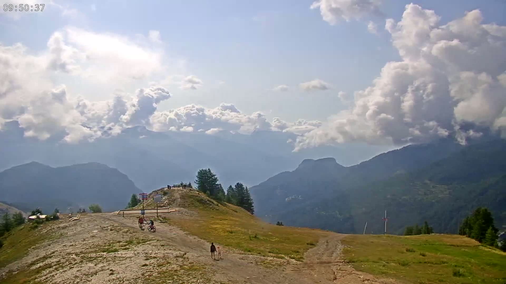 A mountaintop vista shows a partly cloudy sky over a range of distant mountains, with a few people walking and biking on a nearby trail.