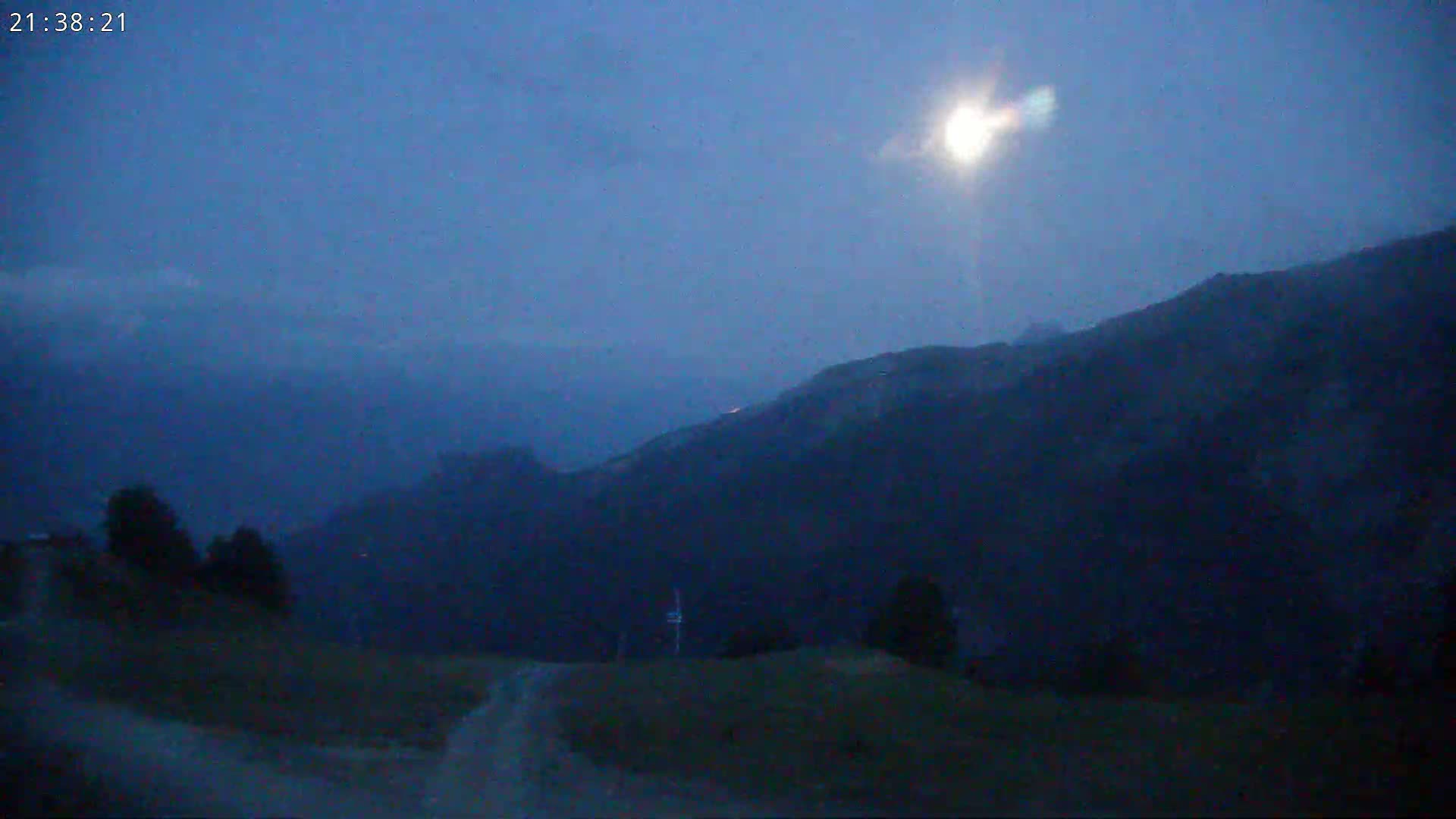 A bright moon shines in a twilight sky over dark, rolling hills and a dirt road.
