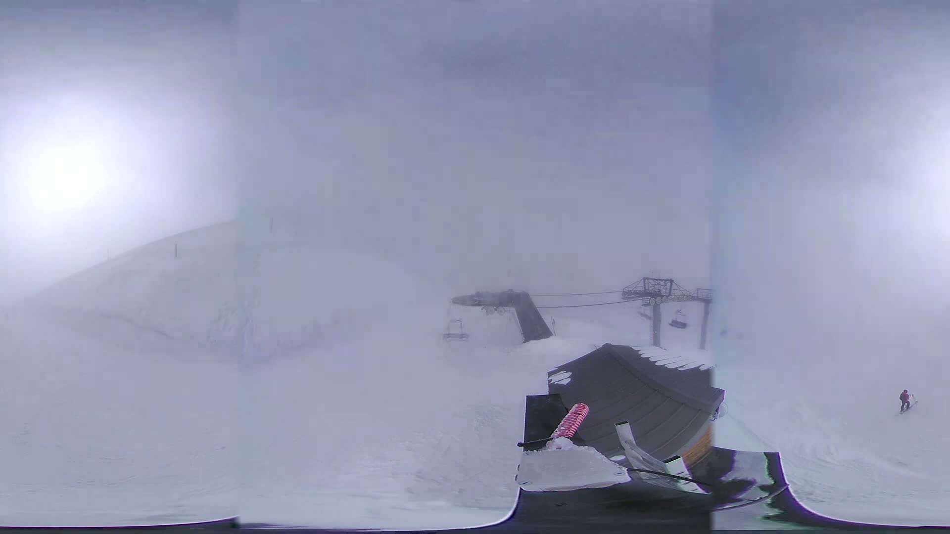 A snow-covered ski resort is shrouded in thick fog, with ski lifts and a lone skier visible in the distance.