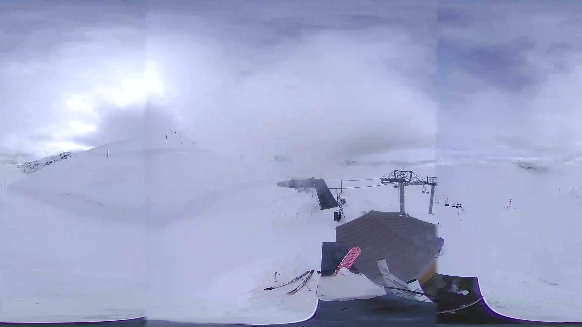 A snowy mountaintop ski resort is shrouded in heavy fog, with a ski lift and a pair of skis visible near a building.