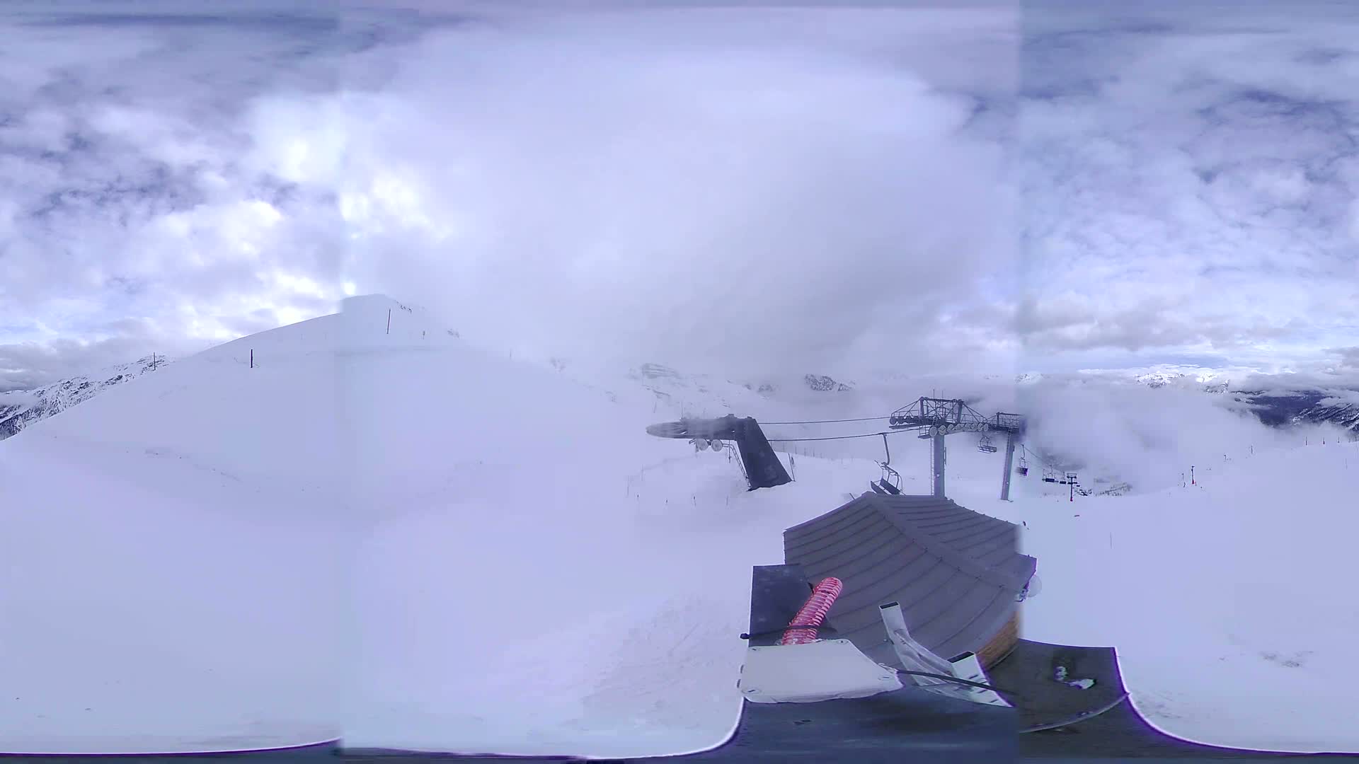 A 360-degree view of a snow-covered mountain slope with a ski lift and a building partially obscured by fog under a partly cloudy sky.