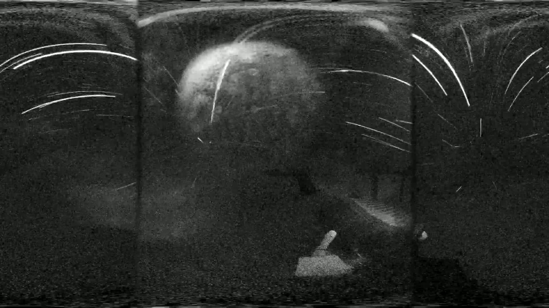 A grainy, black-and-white image shows a large, bright sphere surrounded by numerous streaks of light against a dark background.
