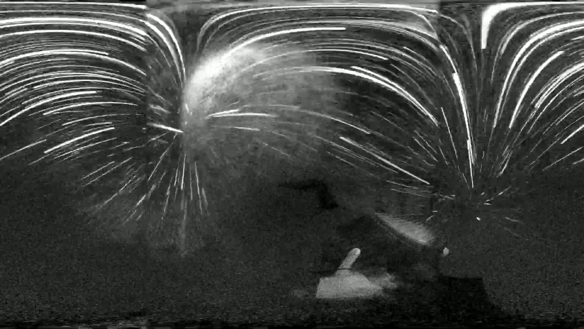 A grayscale image shows a burst of bright lines emanating from a central point, resembling a fireworks display at night.