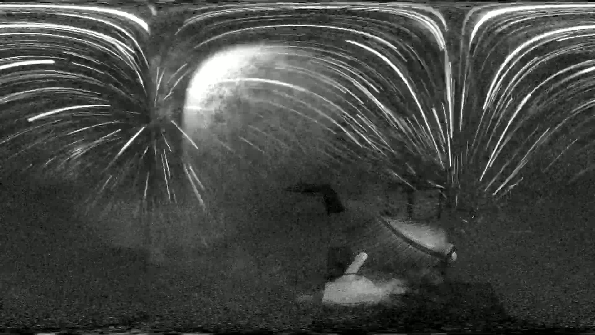 A nighttime, blurry, black and white image shows a bright, full moon with streaks of light radiating outwards, possibly fireworks, behind a dark, indistinct object that appears to be a person or a piece of equipment.
