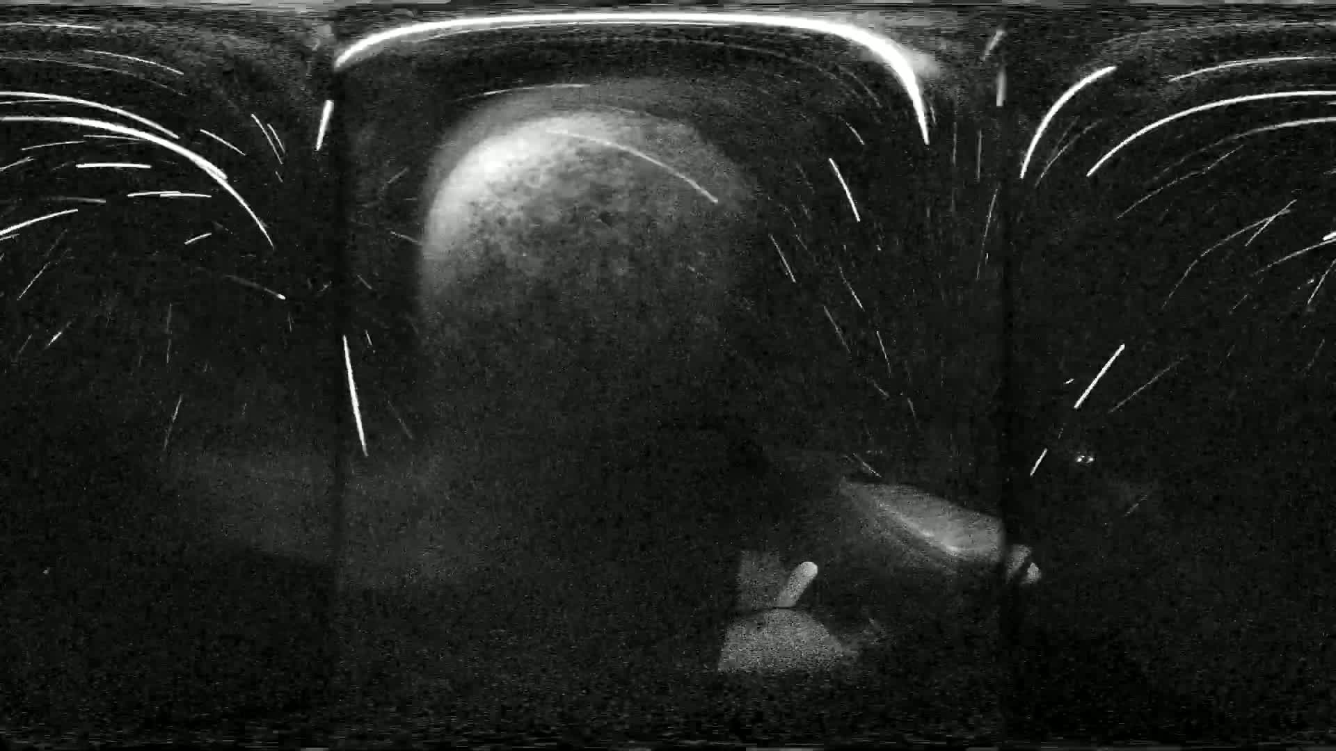 A blurry, dark grayscale image shows streaks of light against a dark background, possibly depicting nighttime, with a large, indistinct object in the center.