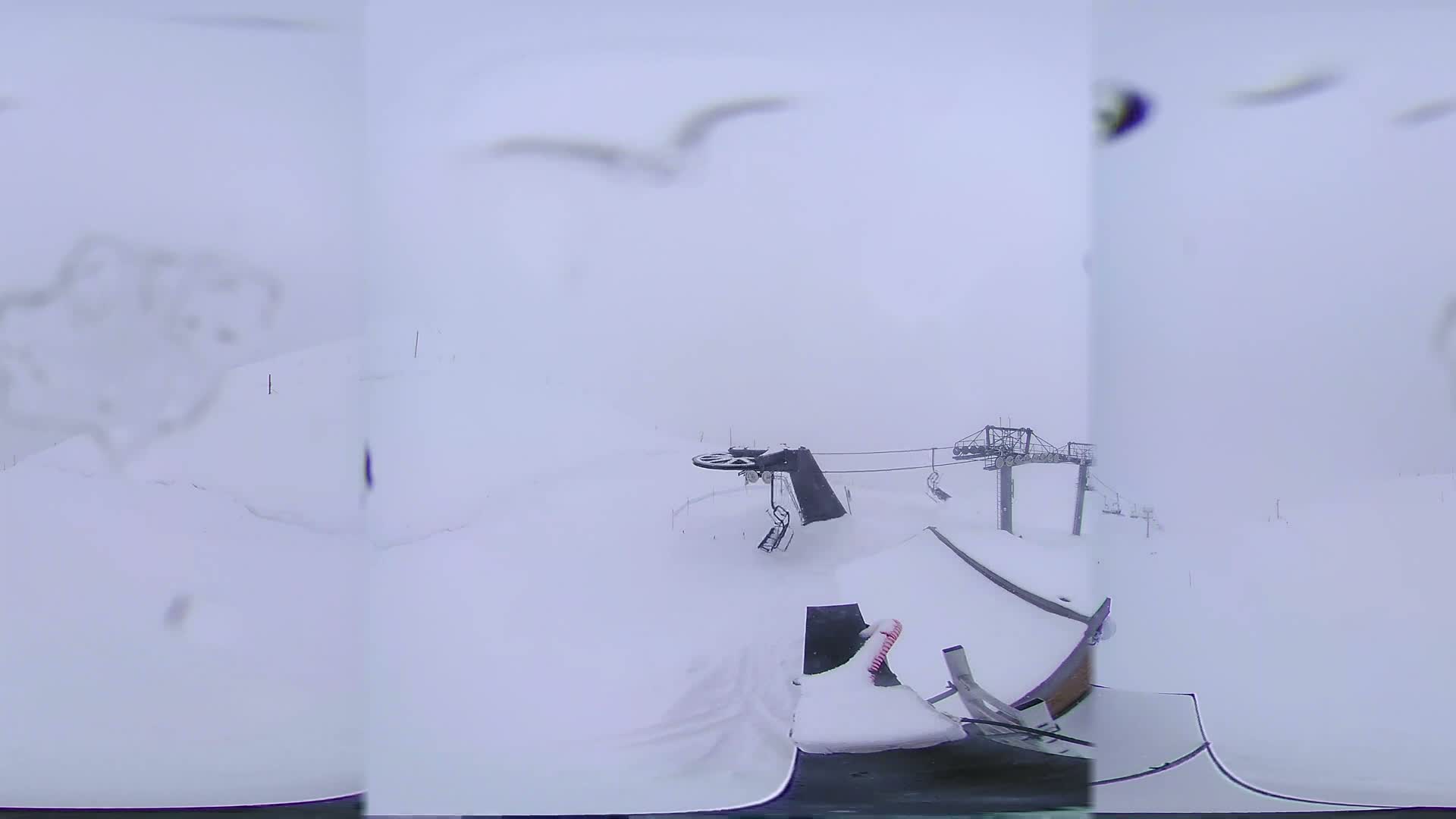 A snow-covered ski lift and surrounding area are visible in foggy conditions with a few birds flying overhead.