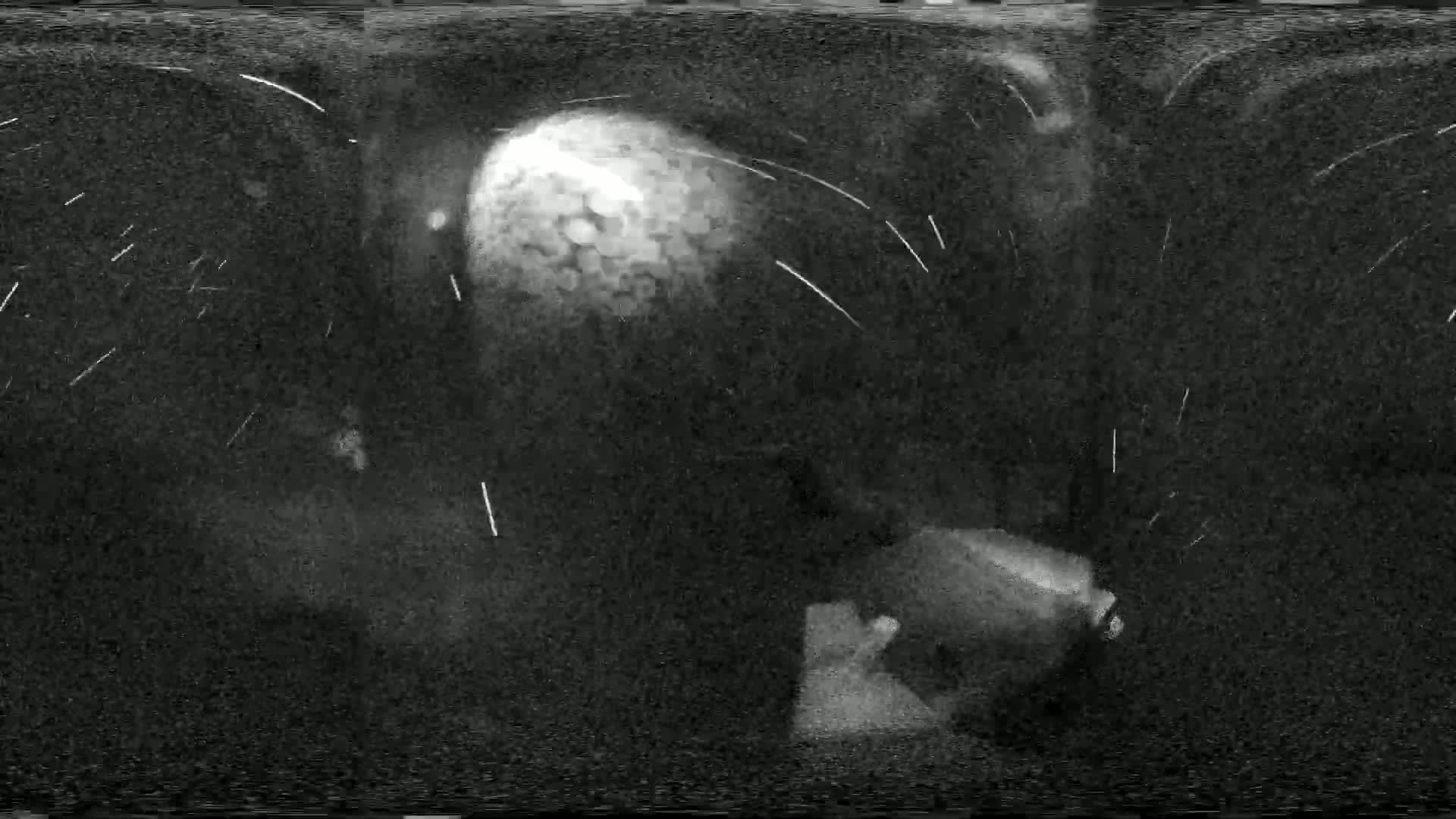 A grainy, black-and-white image shows a large, bright object in the distance, possibly a meteor, with streaks of light suggesting movement through the night sky.