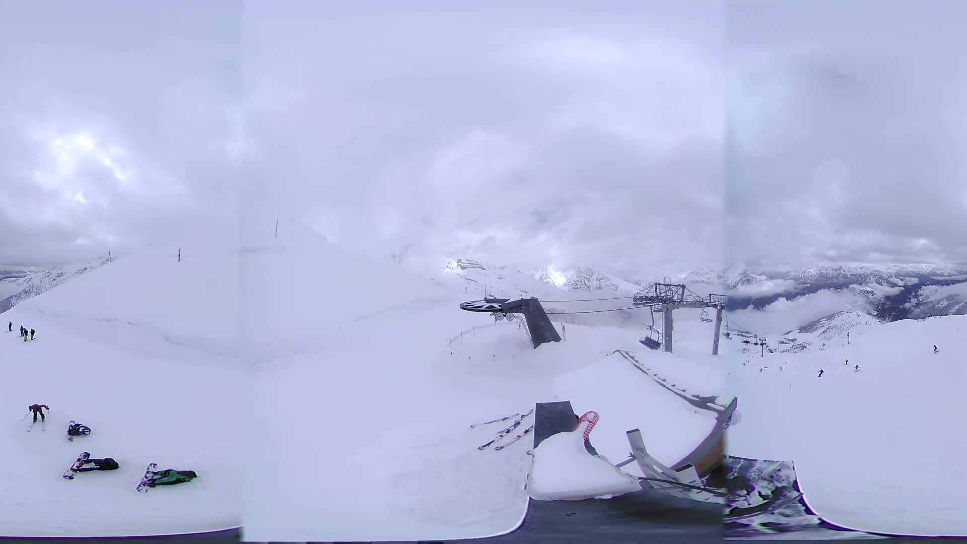 A snowy mountaintop ski resort on an overcast day features a chairlift and several skiers and snowboarders scattered across the slopes.