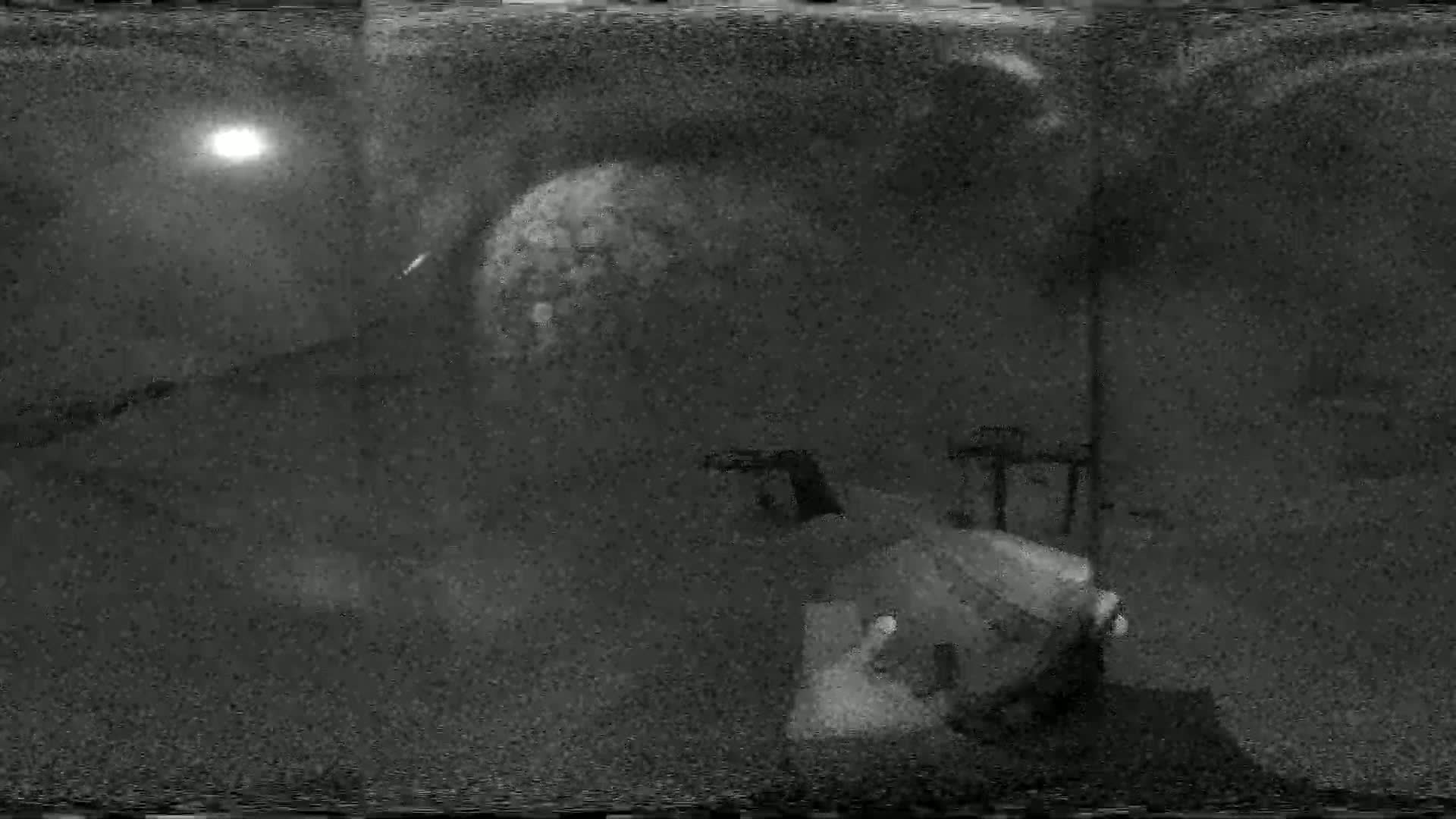 A grainy, low-light image shows a blurry object resembling a large ball in the middle ground, with indistinct structures and a bright light source in the background, under nighttime conditions.