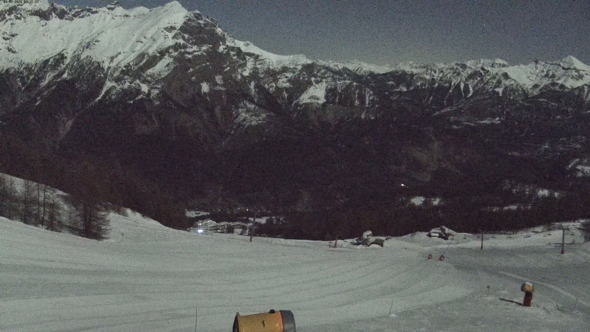 A snow-covered ski slope at night, under a dark sky, overlooks a valley and snow-capped mountains.