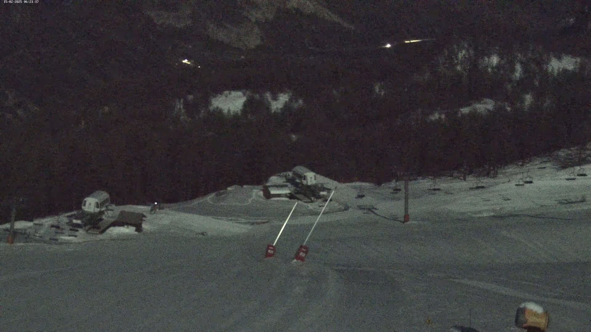 A snow-covered ski slope at night, with a few buildings and ski lift infrastructure visible under dark skies.