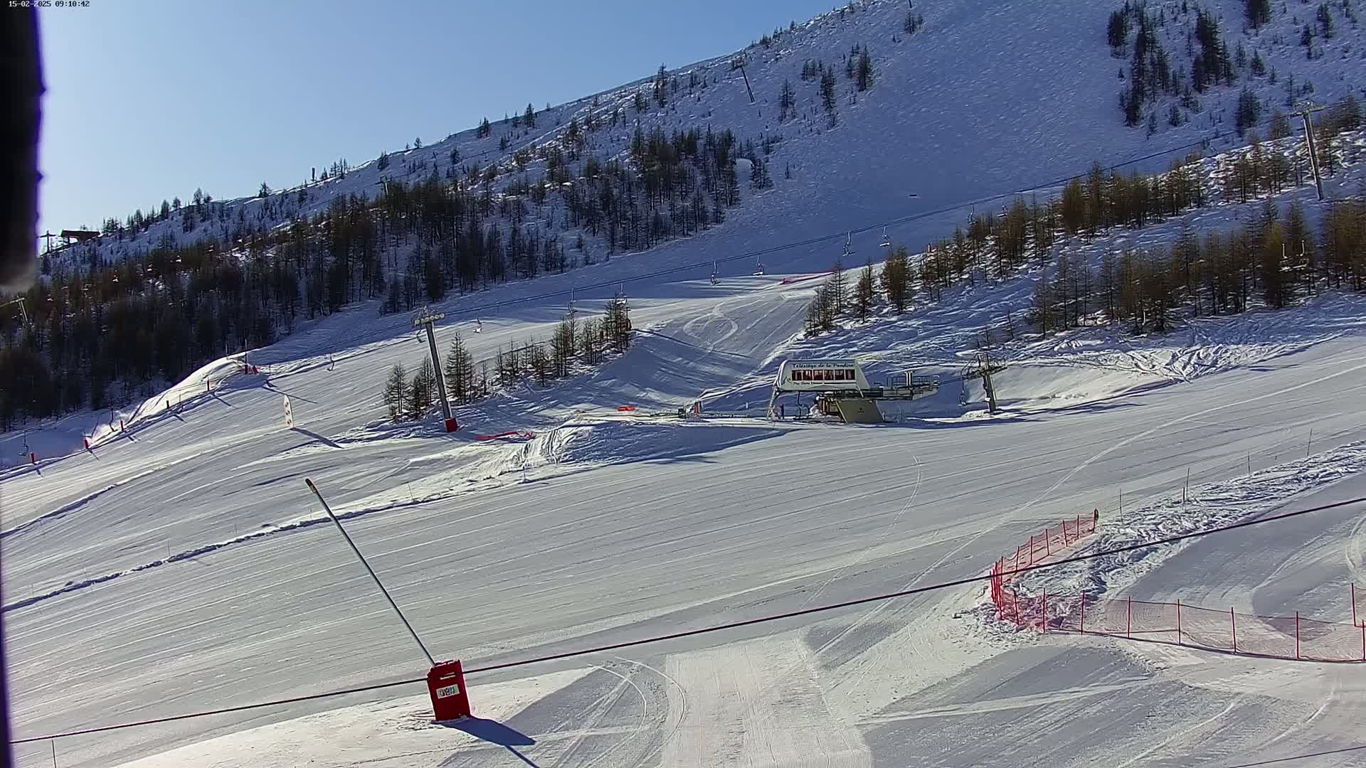 A snow-covered ski slope with several ski lifts and sparsely wooded hillsides under a bright sunny sky.