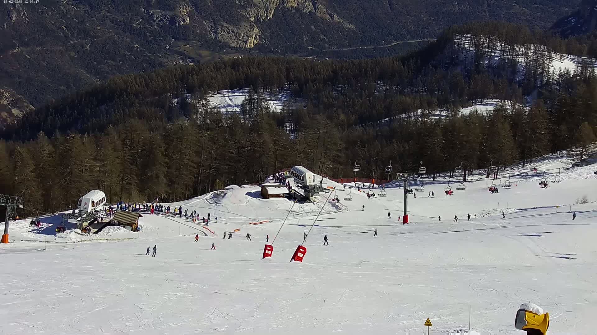 A snowy ski slope with several skiers and snowboarders, chairlifts, and a cable car, is nestled in a mountainous region under a sunny sky.