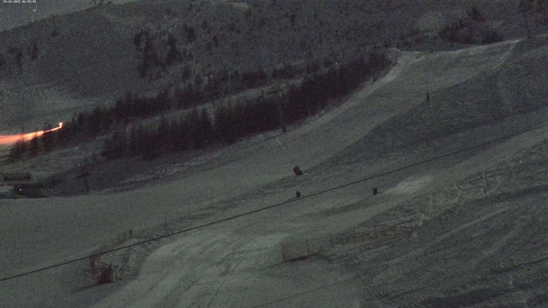 A snow-covered ski slope at twilight, with a few skiers visible and a faint orange glow in the distance.