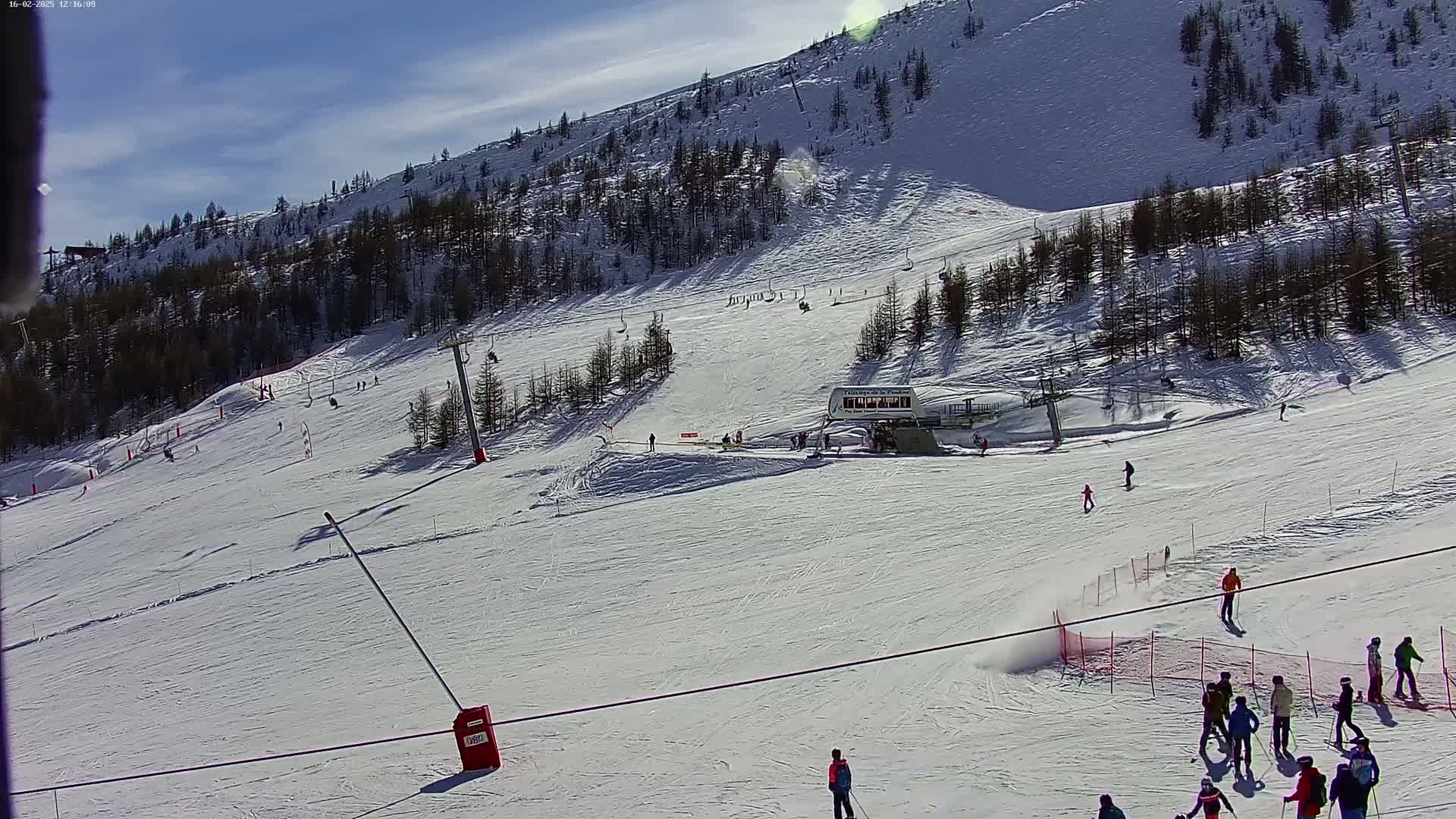 A sunny ski slope with several skiers and snowboarders, a ski lift, and sparse evergreen trees on a snow-covered hillside.