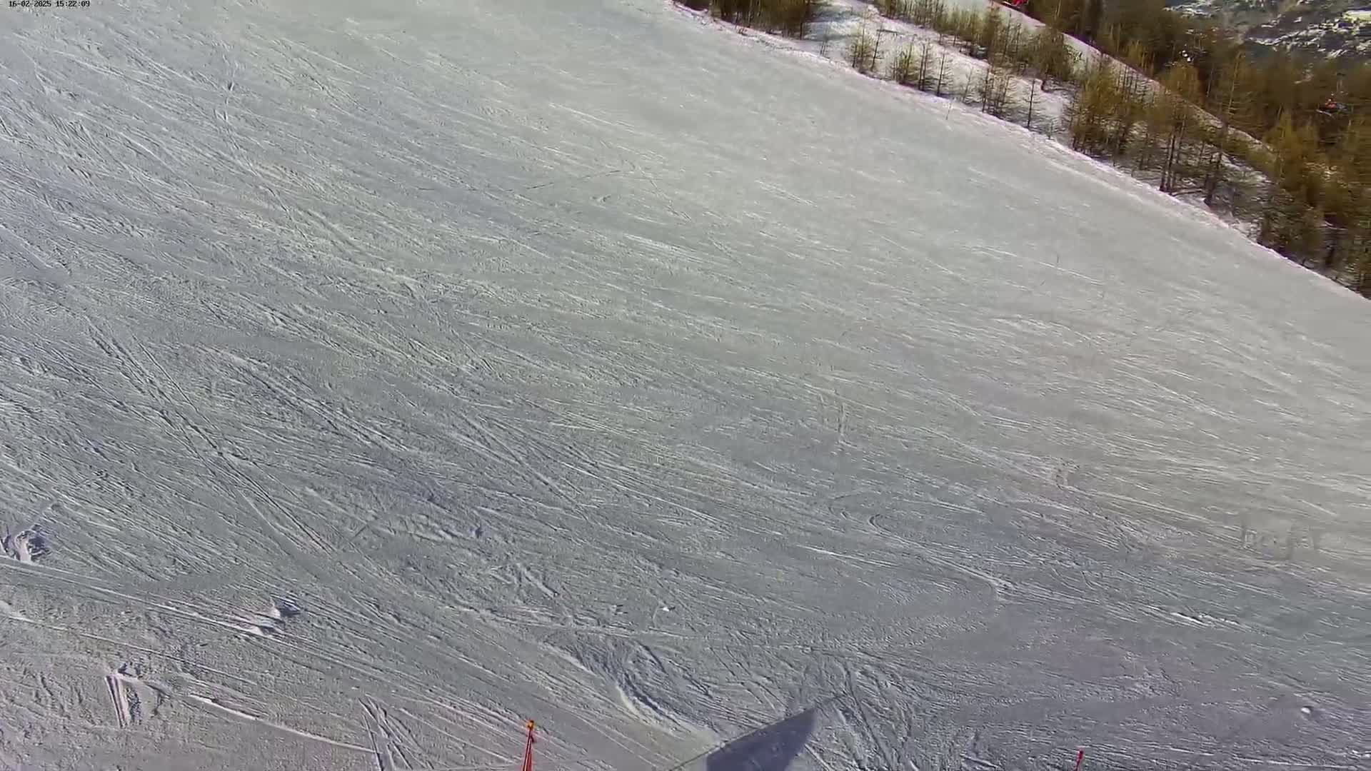 A snow-covered ski slope with numerous ski tracks is seen from an aerial perspective, under bright, sunny conditions.