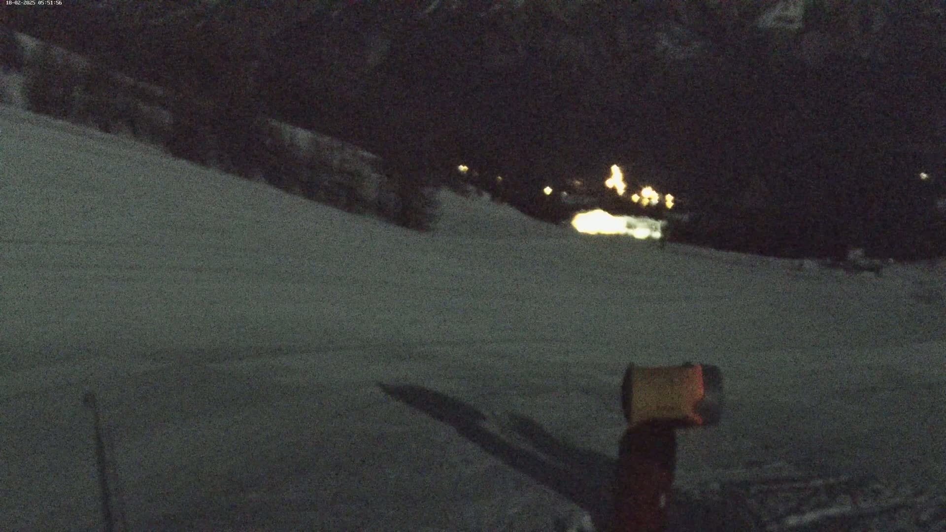 A snow-covered slope at night, illuminated by distant lights, with a snow cannon in the foreground.