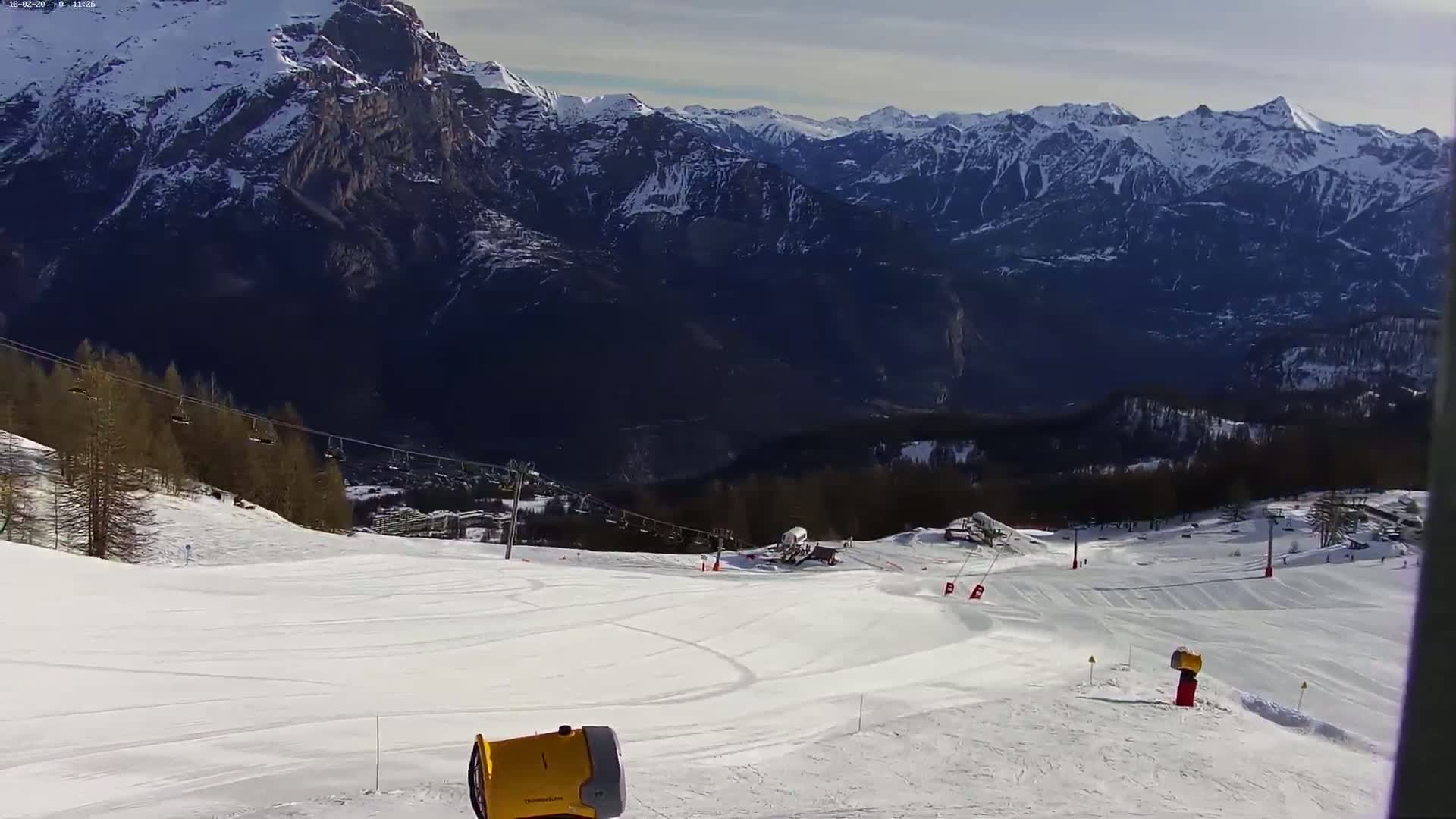 A snowcat is positioned on a snowy ski slope overlooking a valley with snow-capped mountains under a partly sunny sky.