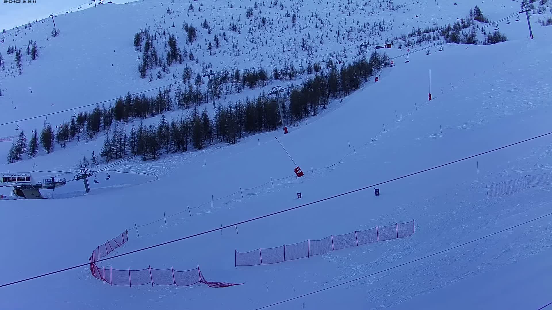 A snow-covered ski slope with a chairlift, evergreen trees, and safety netting under a twilight sky.