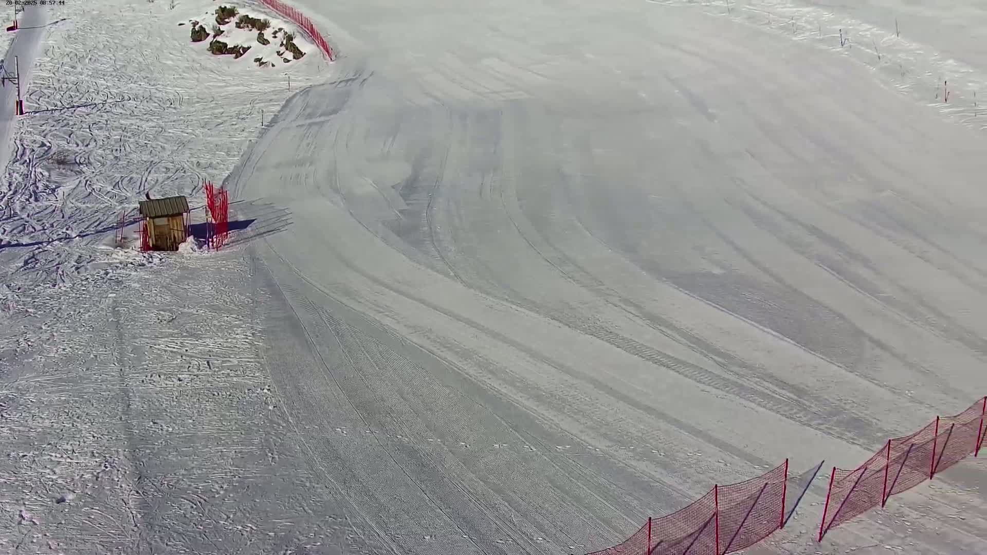An aerial view shows a snow-covered ski slope with tire tracks, a small shed, and red safety nets on a sunny day.