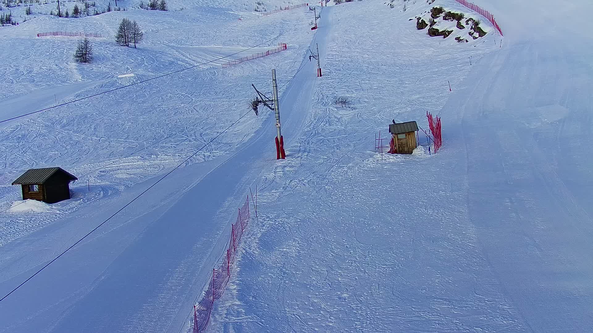 A snow-covered ski slope with a chairlift, two small wooden structures, and red safety netting.