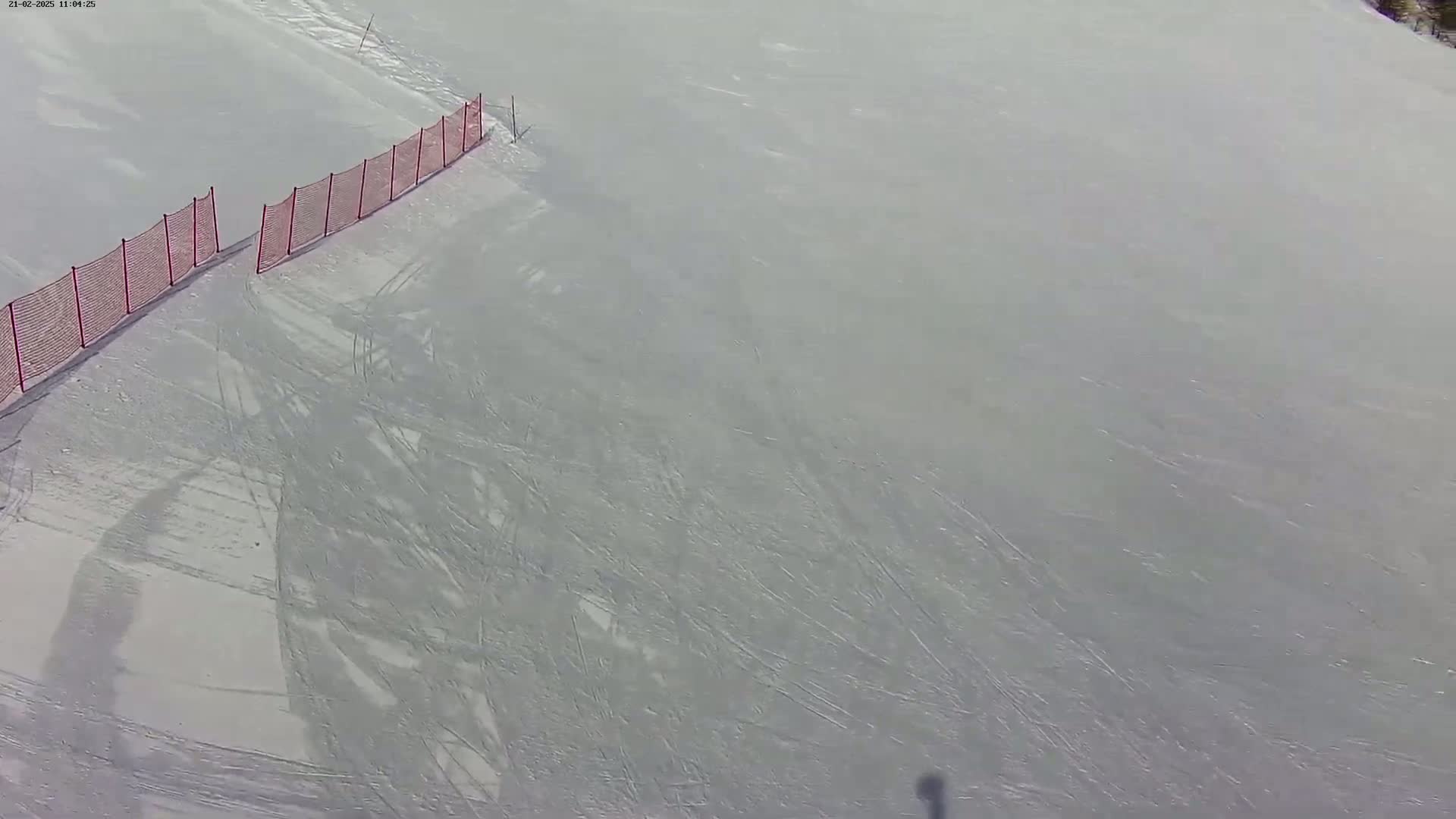 A snow-covered slope shows numerous ski tracks and a partially visible red safety net.