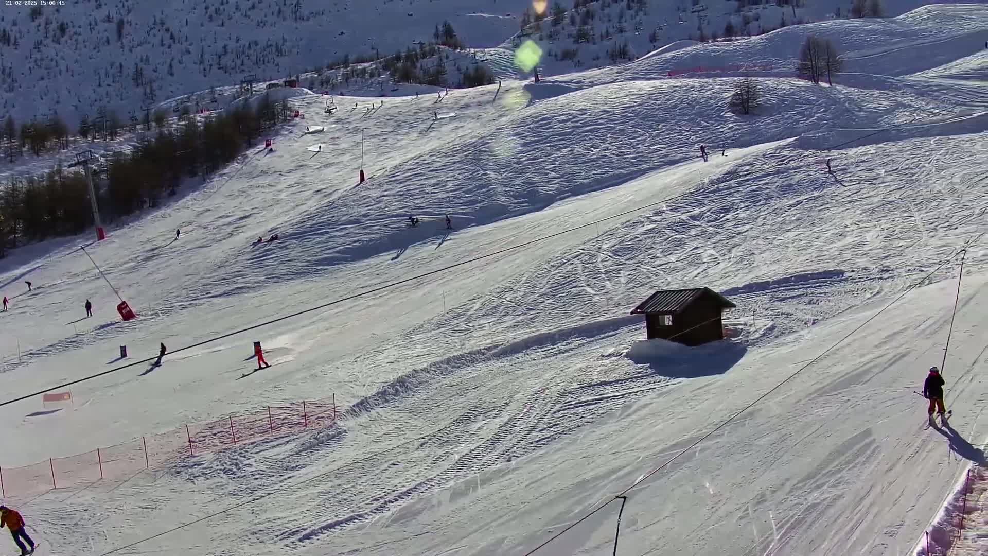 A snowy ski slope with several skiers and snowboarders, a small wooden building, and ski lifts under sunny conditions.