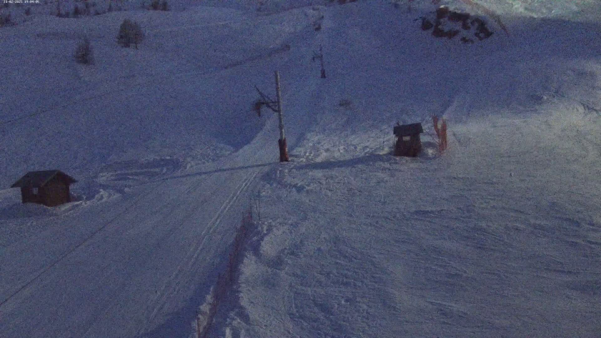 A snow-covered ski slope at twilight, featuring two small structures and a ski lift.