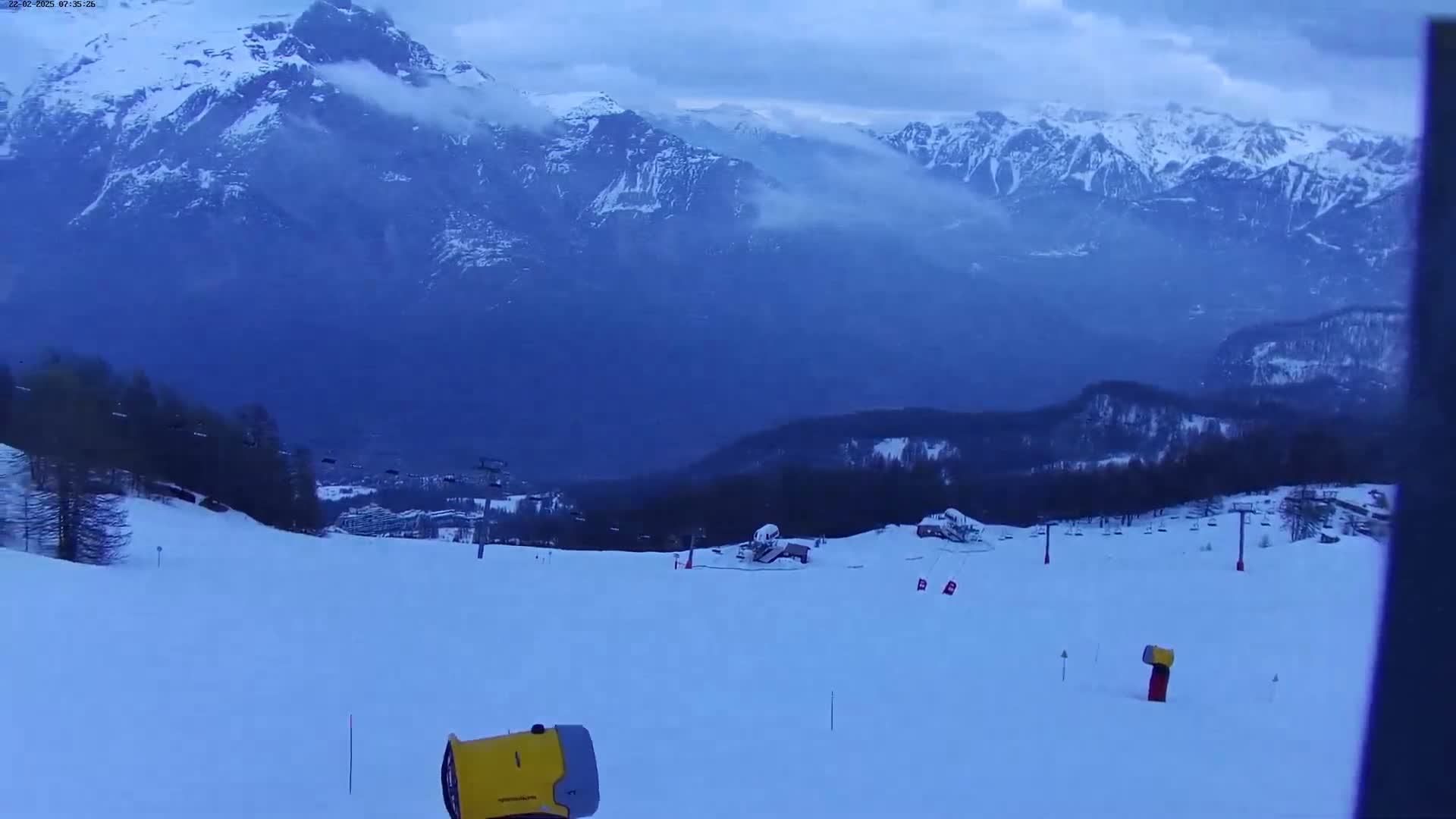 A snow-covered ski slope overlooks a valley and distant, snow-capped mountains under a cloudy sky.