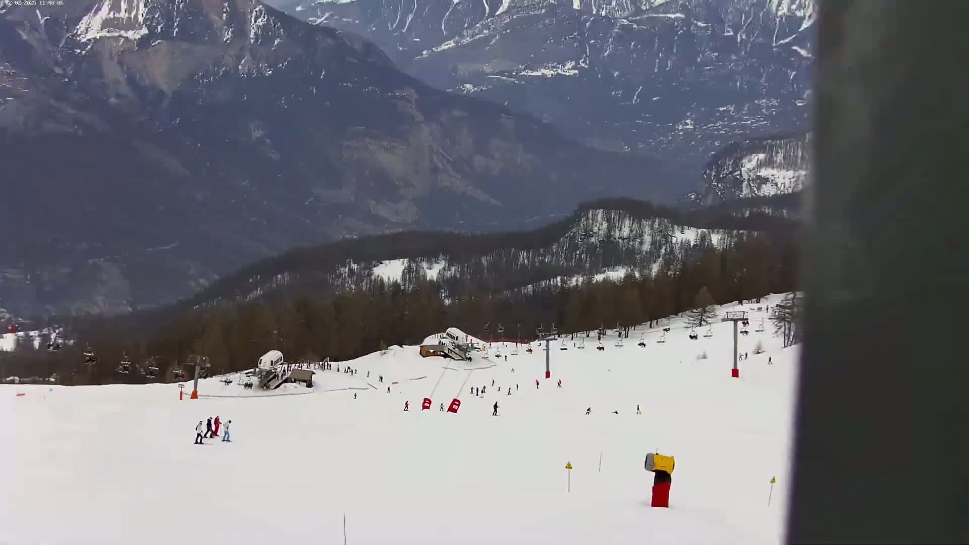 A snowy ski slope with scattered skiers and snowboarders is nestled in a mountainous region under a cloudy sky.
