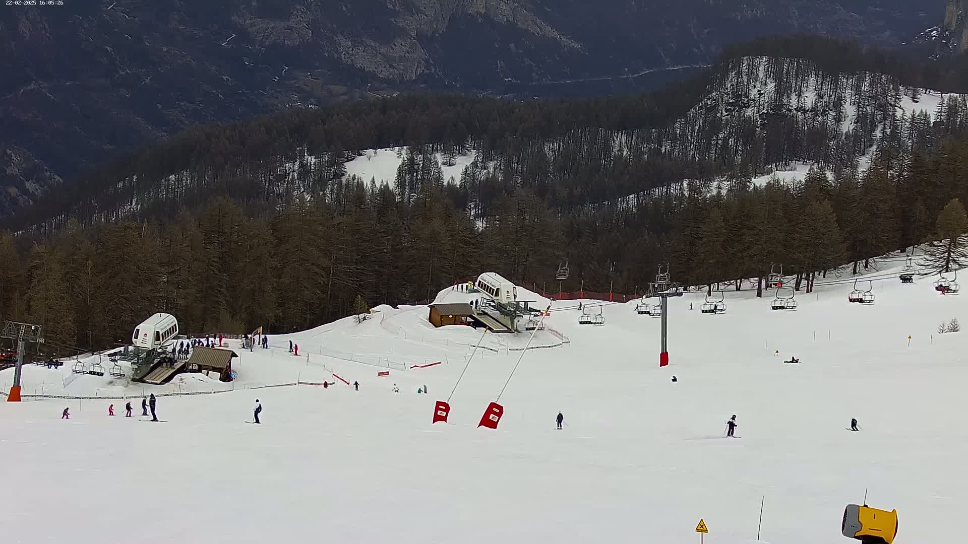 A snowy ski slope with several chairlifts, skiers, and a snow-making machine, set against a backdrop of dark, snow-dusted mountainside trees under a cloudy sky.