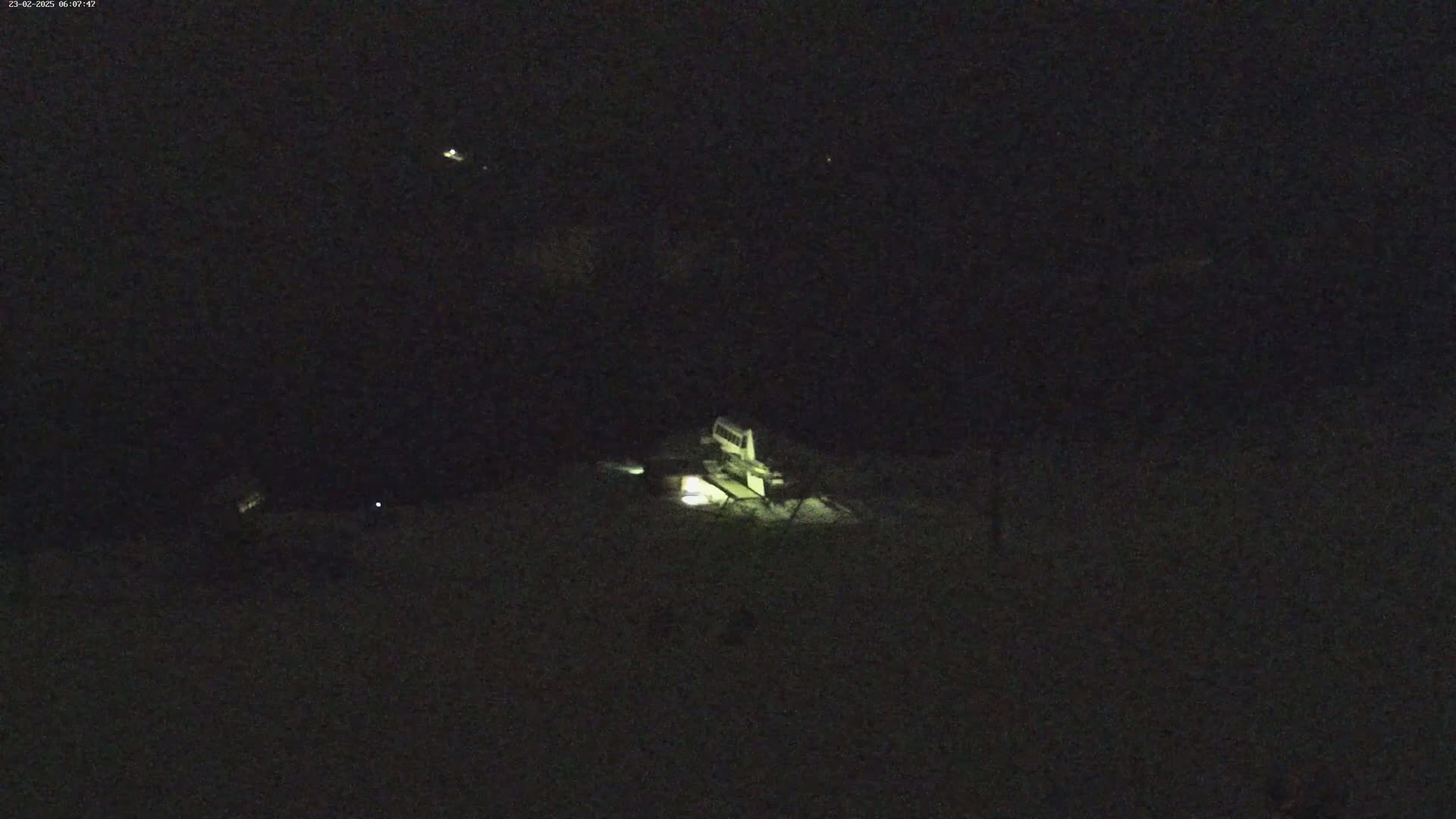 A dimly lit, snow-covered area at night is shown, with a small, illuminated structure in the center.