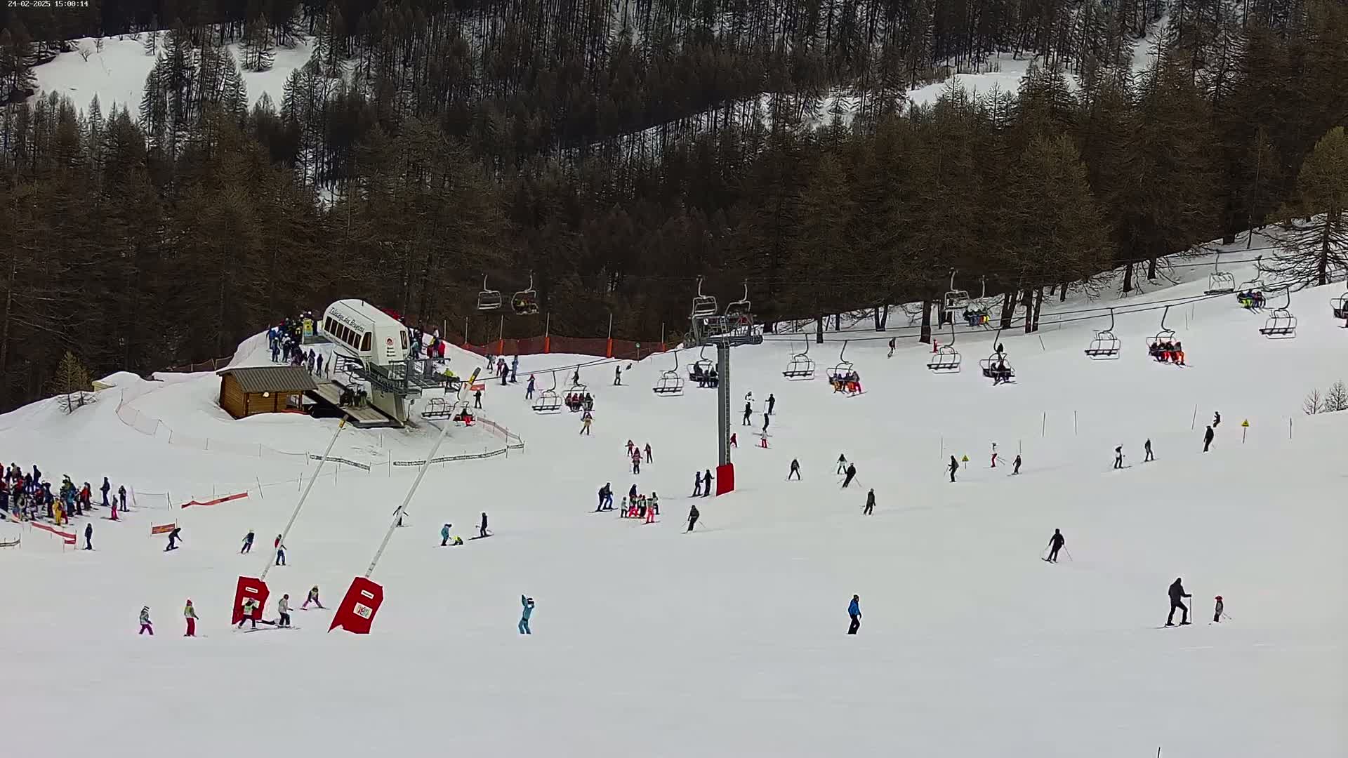 A ski slope with numerous skiers and snowboarders, chairlifts, and a lift building surrounded by a snow-covered forest on a clear day.