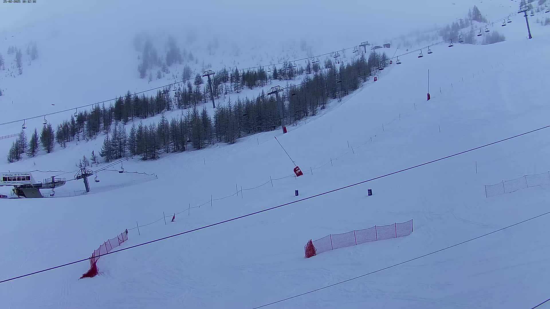 A snow-covered ski slope with chairlifts, sparsely-treed areas, and several orange safety nets under foggy conditions.