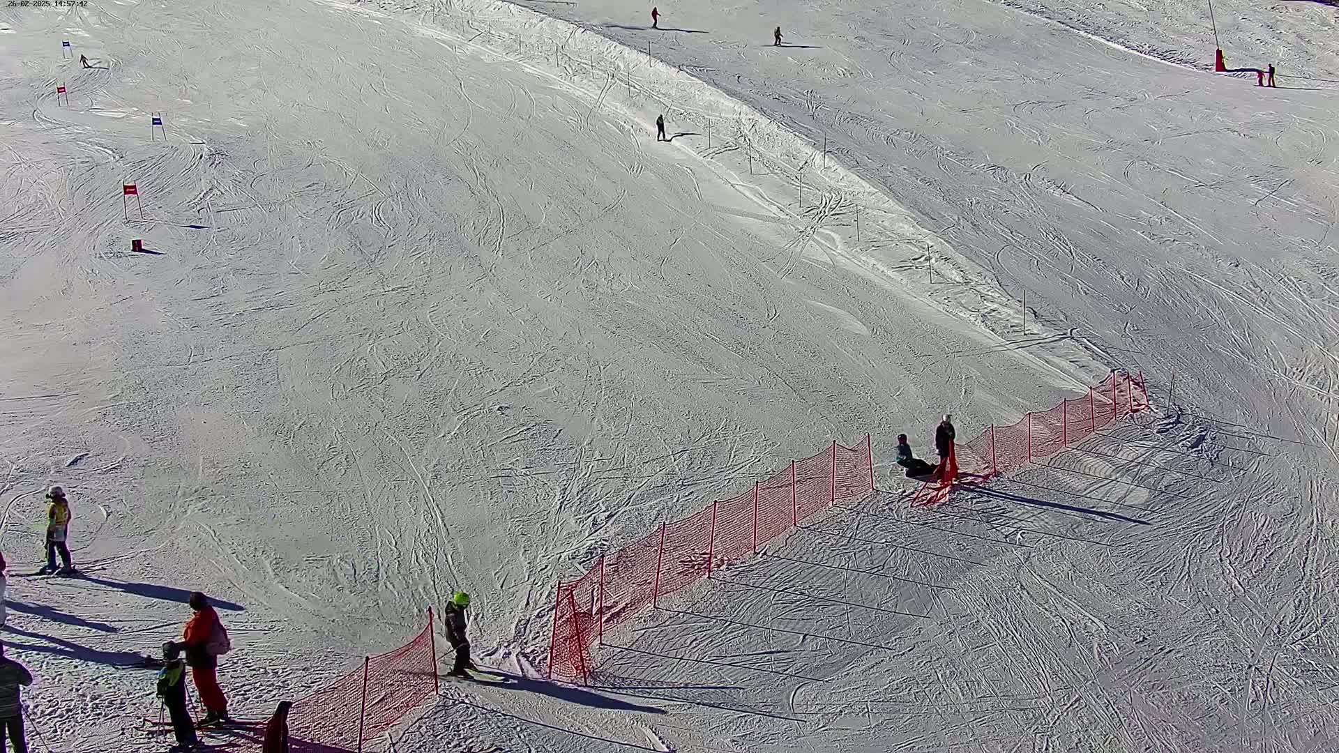 A snowy ski slope with several skiers and snowboarders scattered across it, a safety net along the side, and a sunny day.