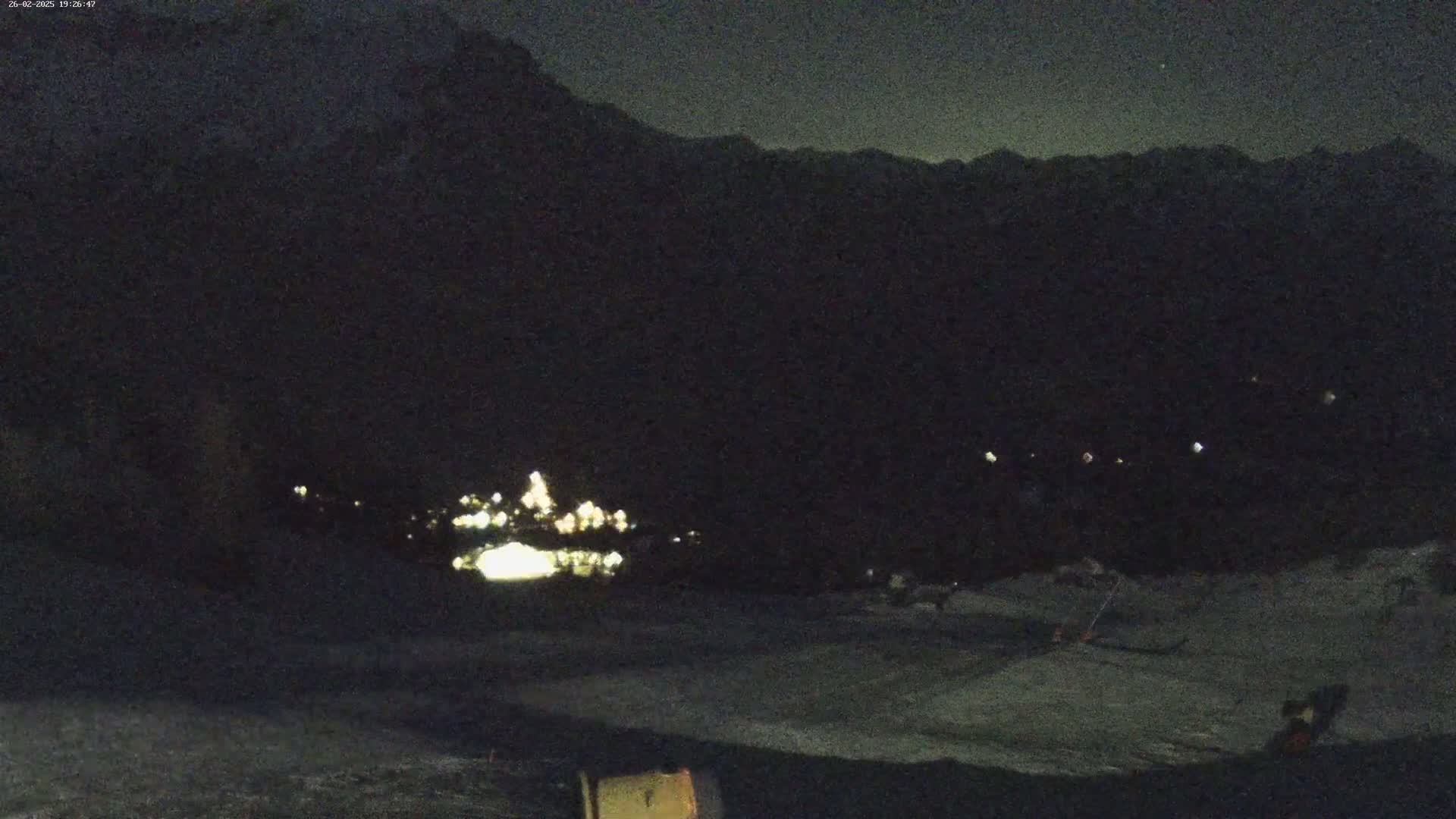 A nighttime view of a town's lights nestled in a valley, with dark mountains rising in the background under a clear night sky.