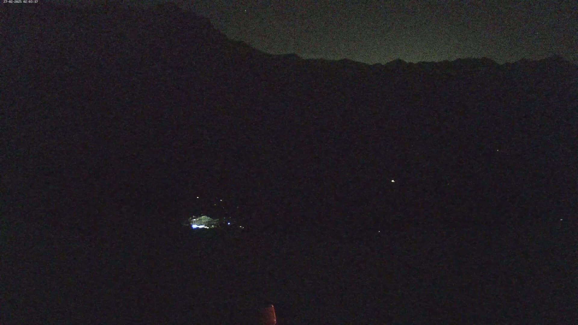 A nighttime long shot shows dimly lit structures below a dark mountain range under a clear, starlit sky.