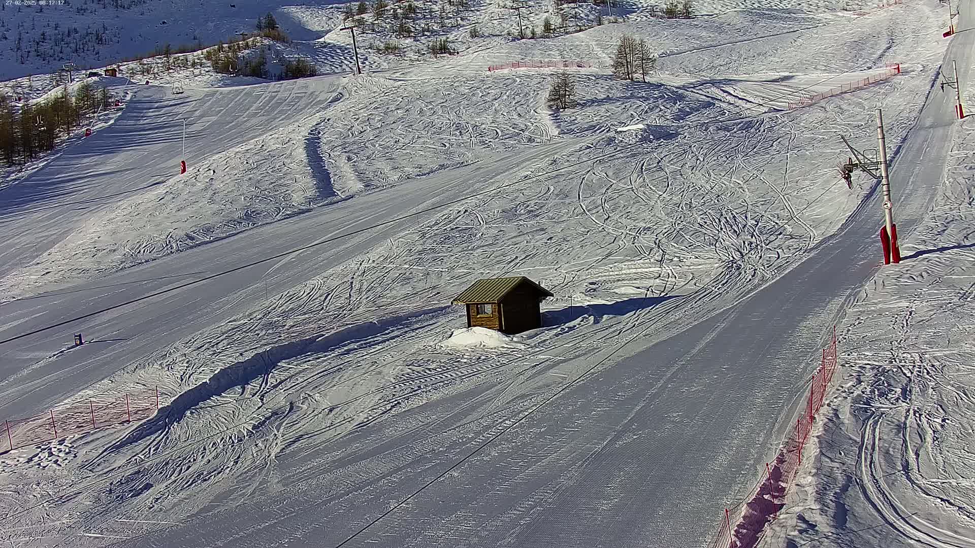 A small wooden cabin sits on a snow-covered ski slope on a sunny day.