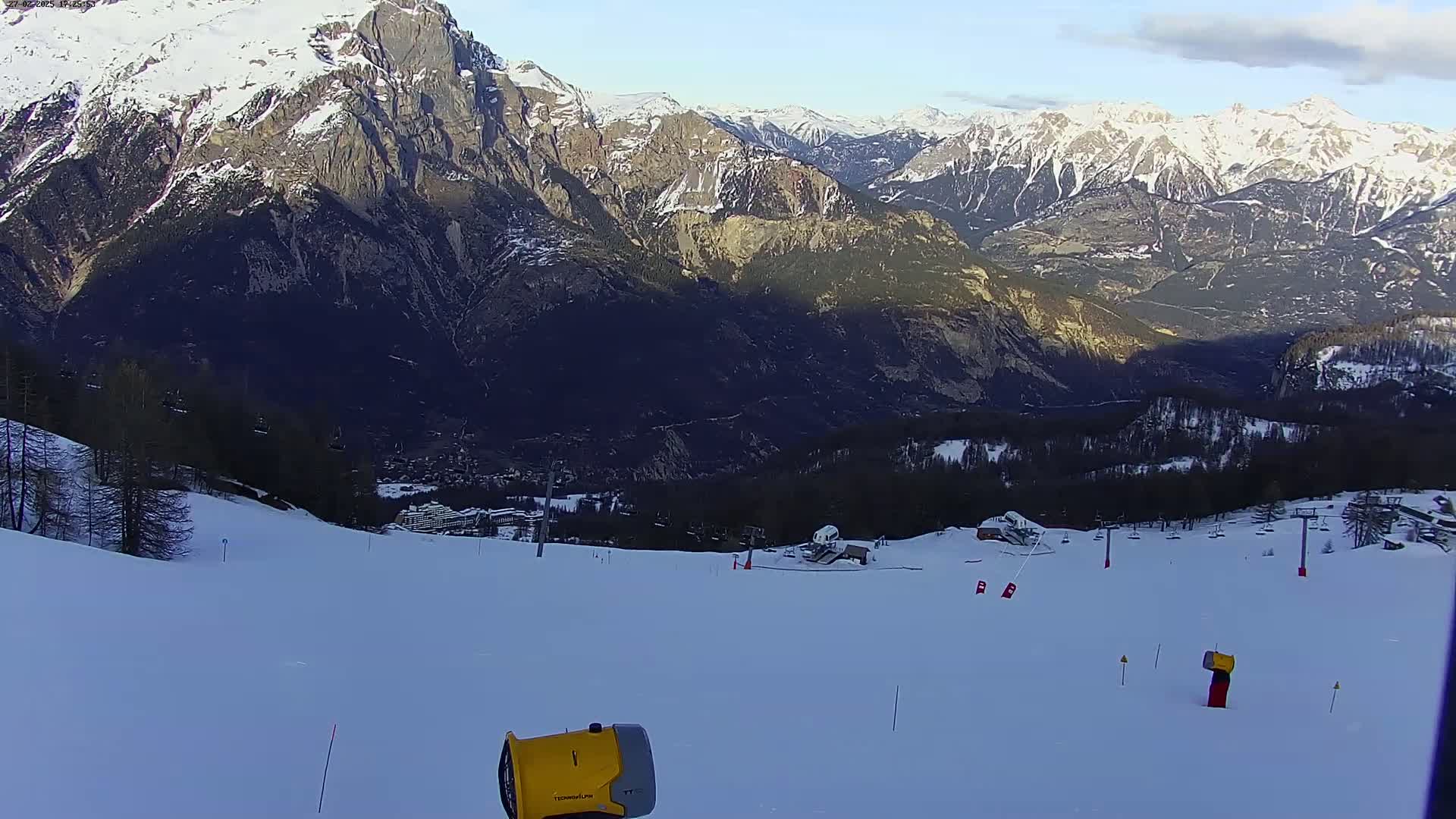 A snow-covered ski slope with a snowmaking machine in the foreground, overlooking a valley and snow-capped mountains under a mostly clear sky.
